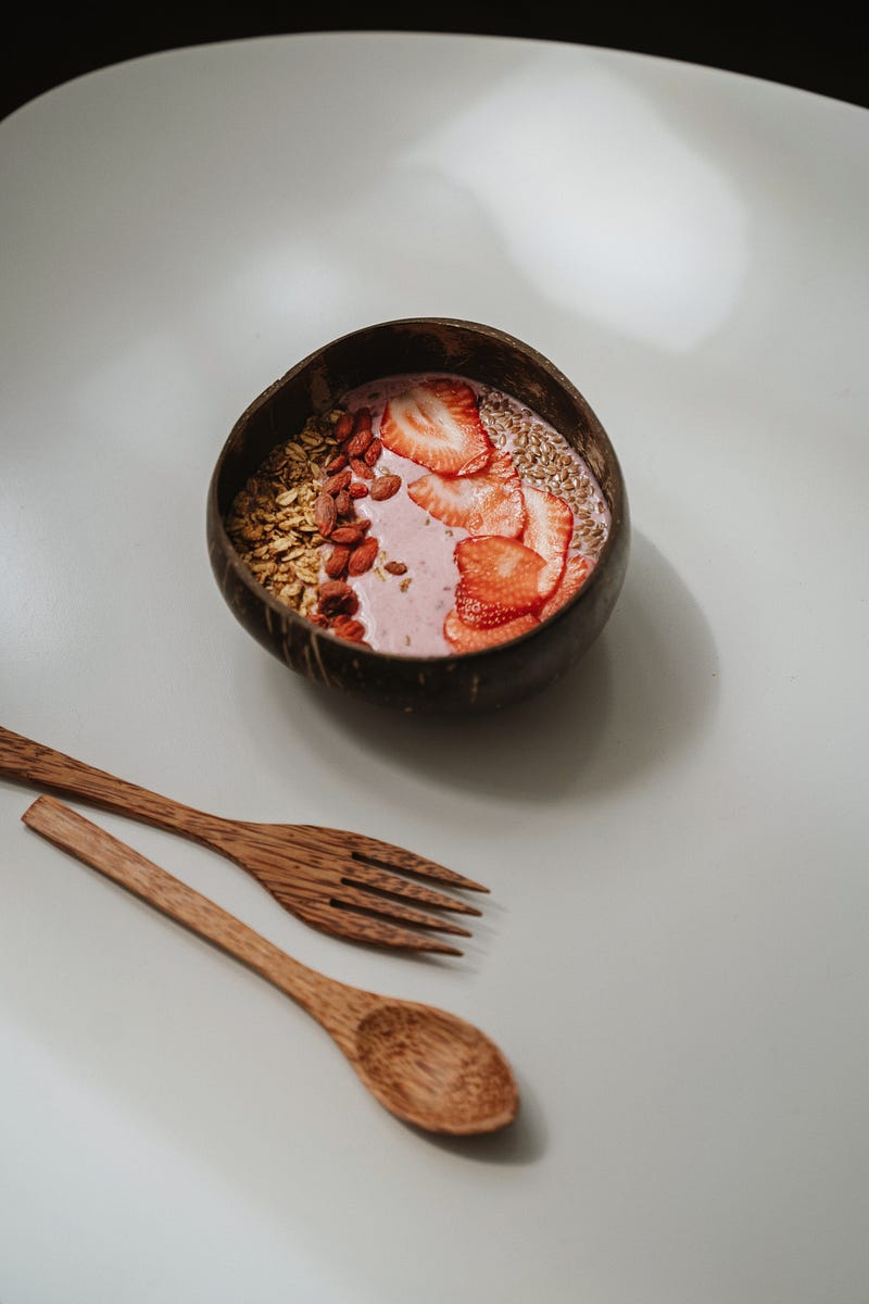 Strawberry smoothie served in a bowl with strawberries and more toppings on top. The bowl along with a wooden spoon and a fork are placed on a white surface.