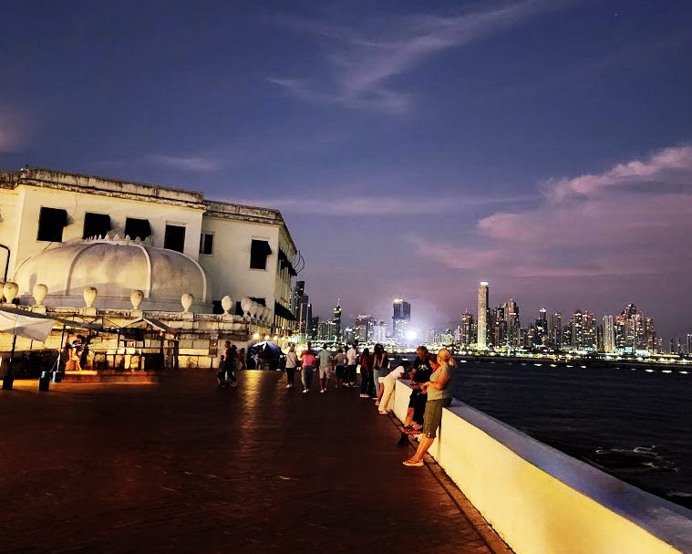 From an oceanfront boardwalk at dusk, in the foreground people pass by an old theater. In the Background the electrified city skyline can be seen.