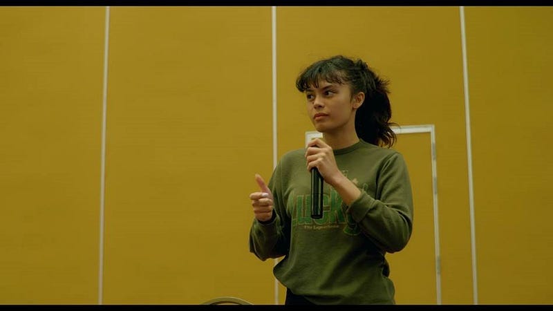 Photo of young woman with dark hair in a ponytail and speaking into a microphone.