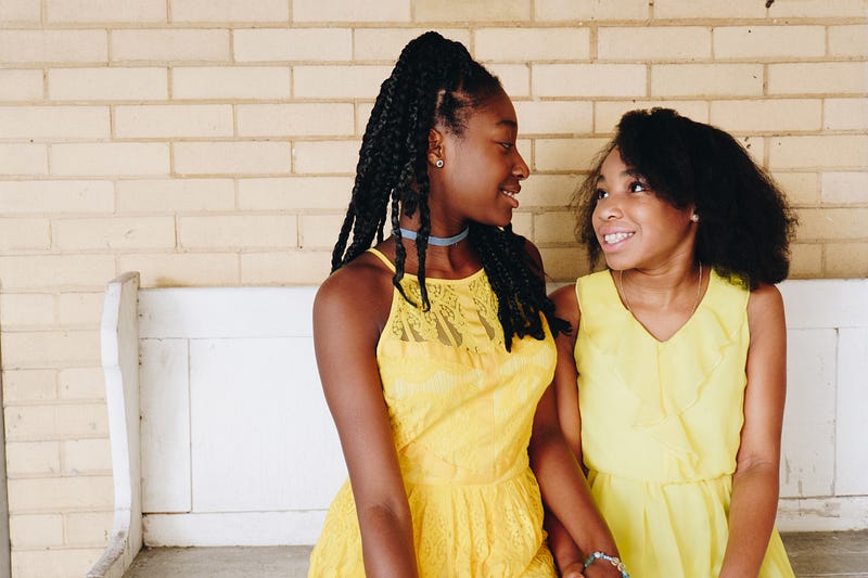 Image of two Black girls wearing yellow dresses looking at each other and smiling.
