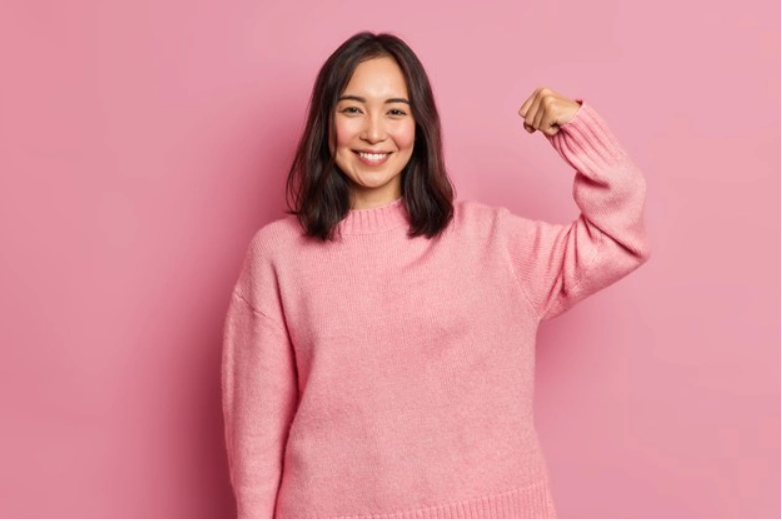 Powerful cheerful brunette woman raises arm and shows muscle demonstrates her strength looks confident at camera smiles gently wears casual long sleeved jumper