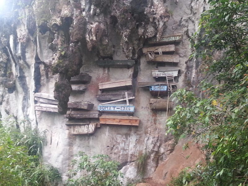 The picture shows a sheer cliff face on which a number of different colored coffins are hanging.