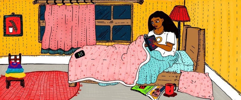 A person with long black hair sits up in bed, under a blanket, in a bedroom, reading a book.