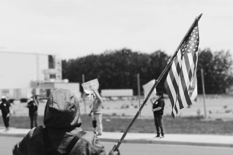 A person wearing a hoodie with the hood up holds a waving American flag. Across the street, protesters stand with signs.