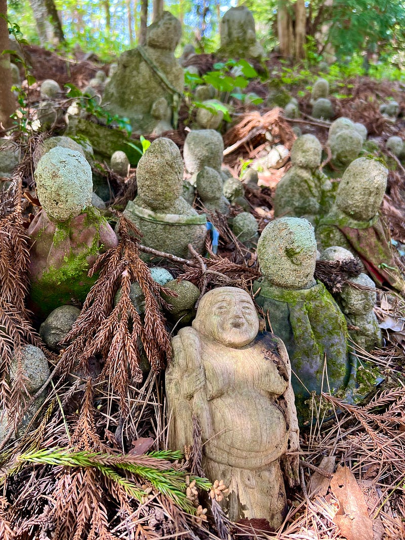 Stone statues and one wooden statue share the ground with pine needles at the Nashi no Ki Jizō.