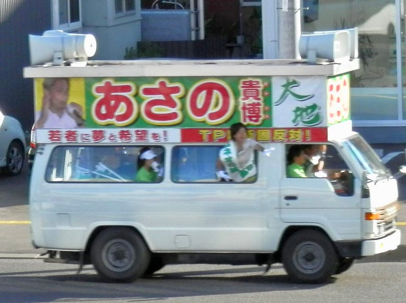 Van equipped with large signs advertising a political candidate, speakers on the front and back, and people waving out the windows.
