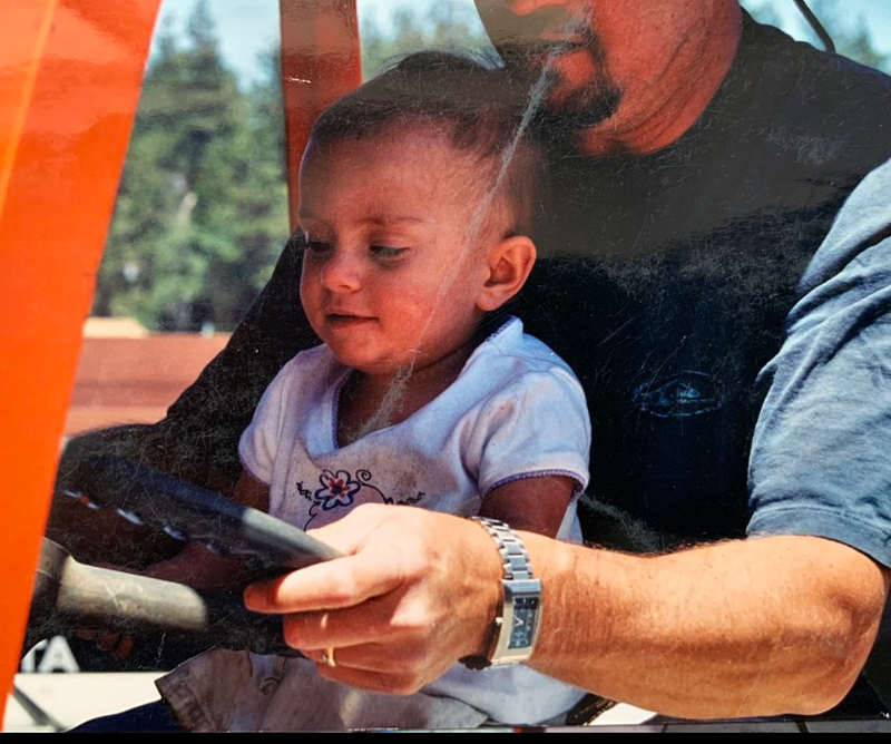 An image of me at 3, on my dad’s lap, driving an orange forklift with trees in the background.