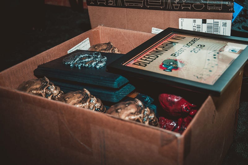 A cardboard box holds a handful of gold-painted skulls and one red-painted skull. The skulls have unrealistic but horrific features. A couple of plaques with flat versions of the skull in silver sit on top of the 3D skulls and balanced on top of the box is a framed certificate reading, “Bleedingham, best score 2018.”