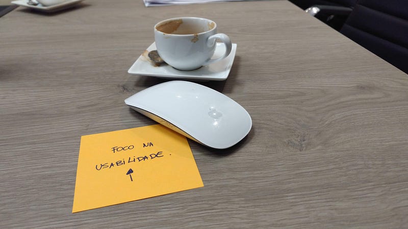 Sobre uma mesa de madeira, uma xícara branca que mostra que um café expresso foi tomado há pouco. À sua frente, um Magic Mouse, da Apple. Sob ele, um post-it cor de laranja exibe o texto "foco na usabilidade" acima de uma seta que aponta para o alto.
