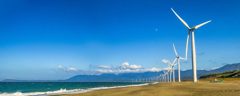 On the right side of the photo, 20 windmills are lined up along the coastline. In contrast, the other side of the photo shows the coastline meeting the ocean, the sky, and the mountains on the horizon. An almost full moon is peeking in above the windmills.