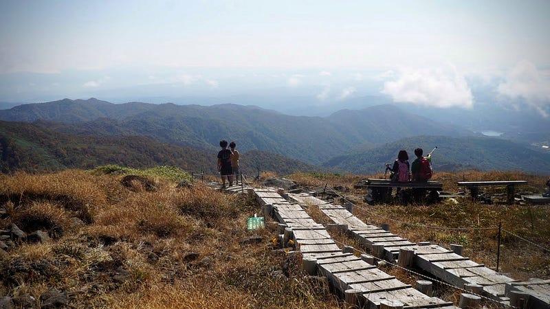 A couple take a rest and point out over to the far hills from a lookout on Mt. Ubagatake, with another couple standing on the wooden path the leads all the way up the mountain
