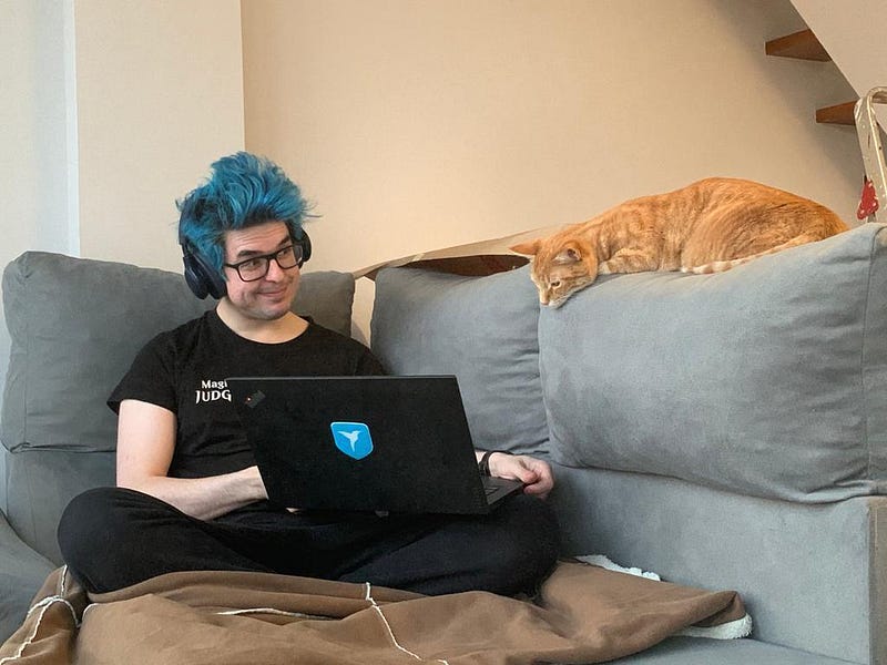 A person in a black t-shirt on a blue couch shows the screen of the laptop he is holding to an orange cat perched on one of the pillows, as if it is can read the screen.