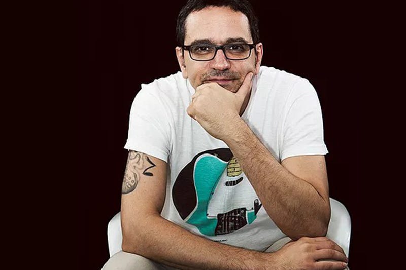 Foto do palestrante Joni Galvão. Ele está sentado, com uma mão no joelho e outra no queixo.