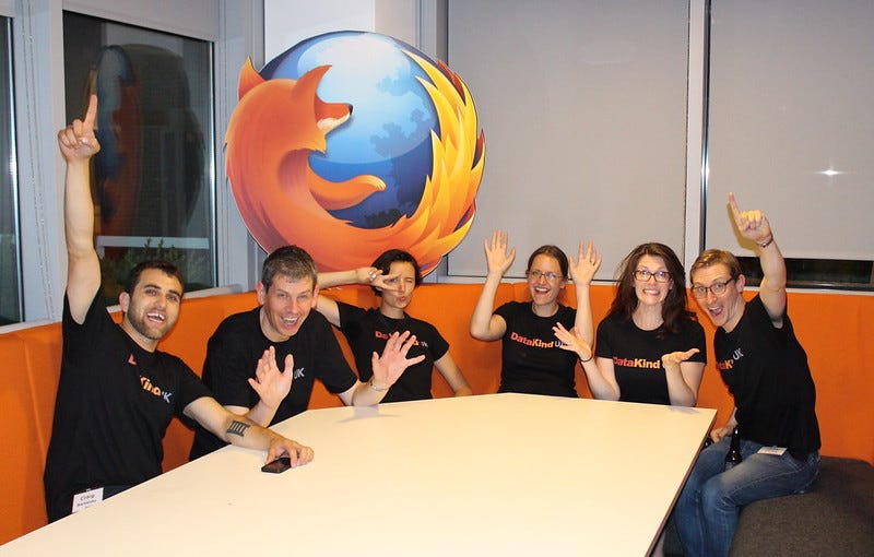 Group image of people at table, smiling and waving, wearing t-shirts that say ‘DataKind’