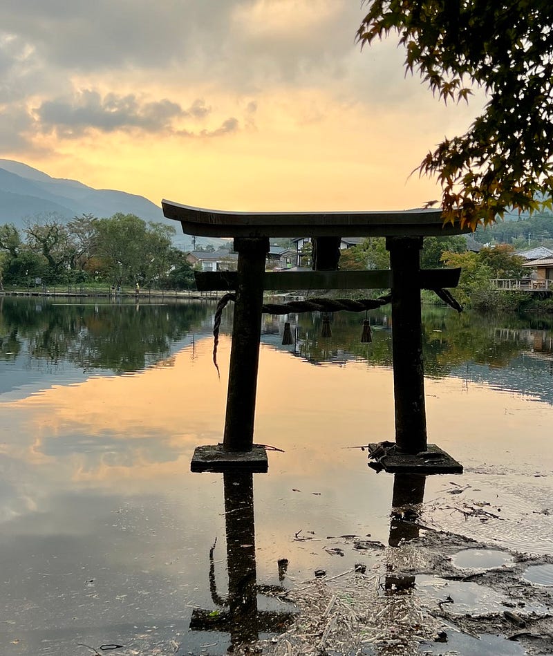 Torii gate rising from a still pond reflecting the surrounding mountains and sky at sunset.