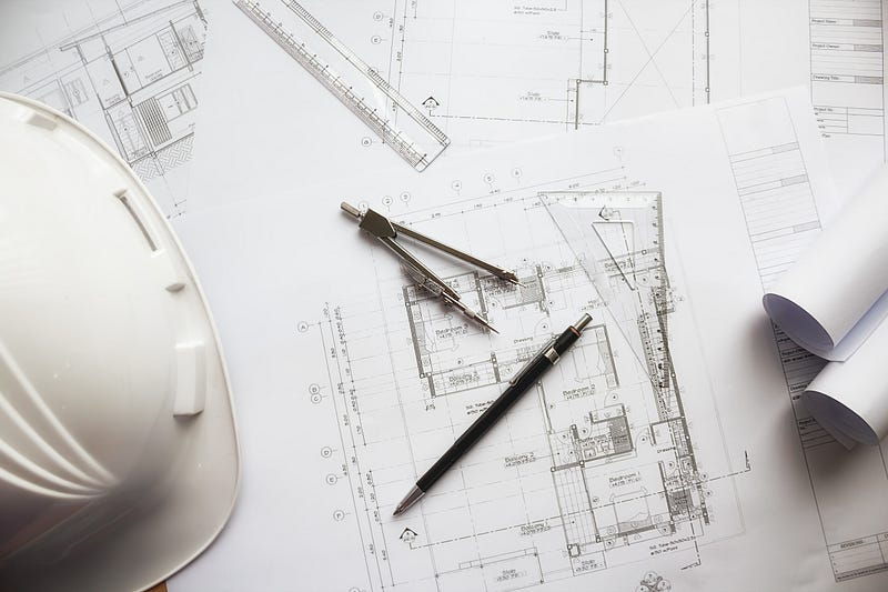 This image showcases a detailed architectural drawing with precision tools, including a compass, scale ruler, and pen placed over residential floor plans. A safety helmet highlights the connection between design and on-site execution. It represents the meticulous planning, technical accuracy, and professional approach required in modern architecture and construction projects, ensuring structurally sound and well-planned spaces.