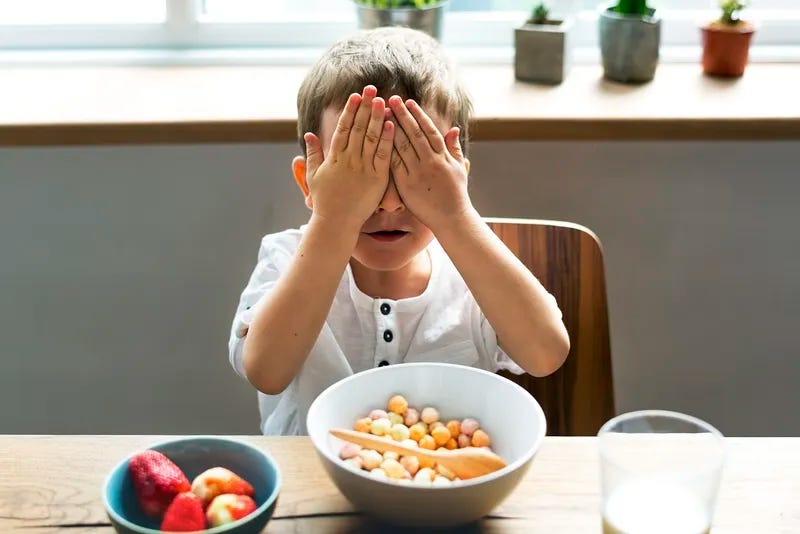 A small boy has his eyes closed with his hands and a bowl of cereal is before him