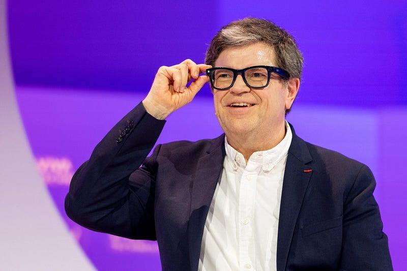 Yann LeCun smiling on stage, wearing black-framed Meta Al Glasses and a dark blazer against a purple-lit background.