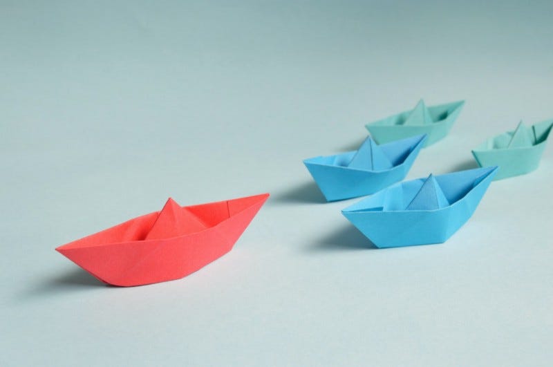 barcos de papel, um na cor vermelha posicionado na frente e 4 barcos atrás, onde 2 são com tom de azul claro e 2 são com tom verde claro
