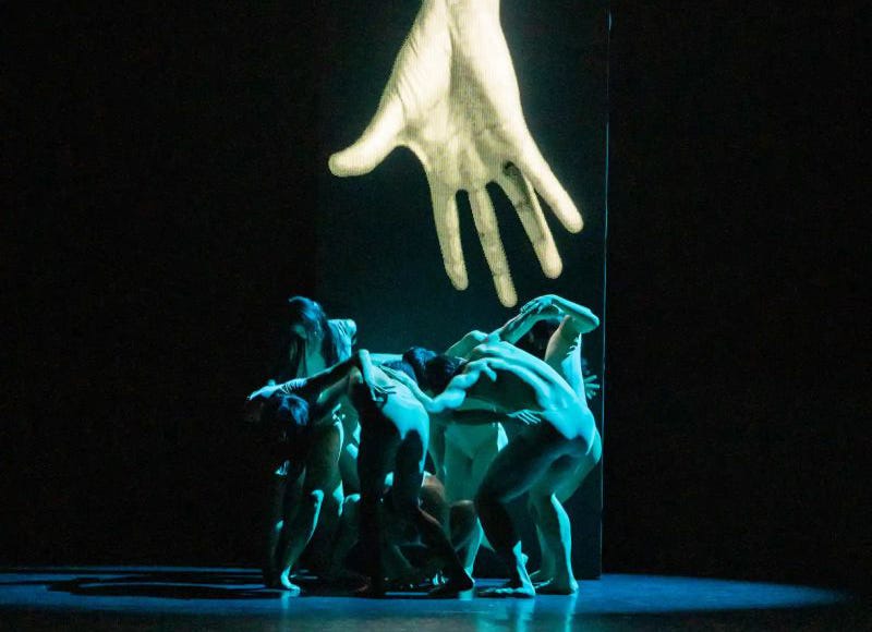 In a dimly lit theater, dancers cluster together under a large projection of a hand, creating a poignant tableau of human connection against a backdrop of digital imagery.