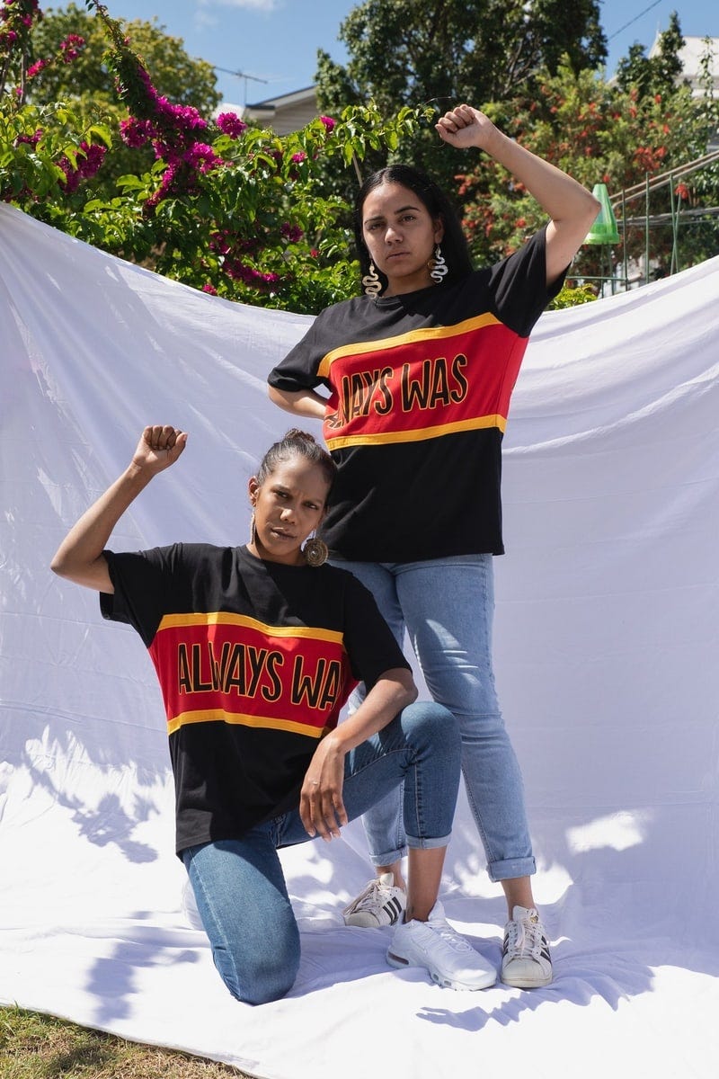 Two people in black shirts with a stripe across the chest. The stripe is red with a yellow outline on either side, and reads ‘Always Was’ in black text. One person is standing while the other is kneeling in front of them. Both have a fist raised.