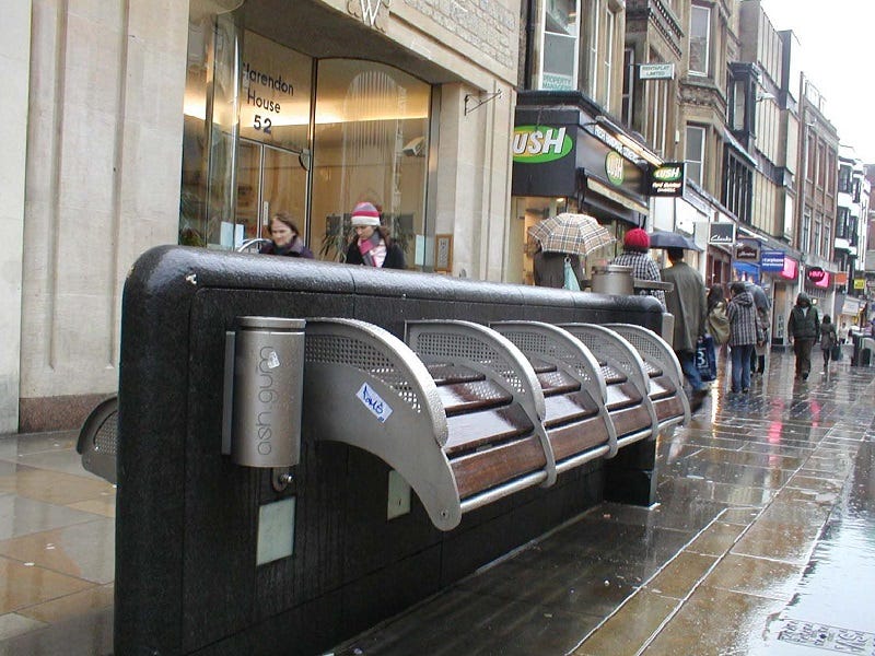 An image of a bank of “benches” on a rainy street. The “benches” slope downward at an angle specifically designed to prevent anyone sitting down on them. Rather, people can only lean against them and effectively remain standing. These are described as “anti-loiter benches.” In the background, people can be seen walking between different stores on a busy shopping street in a city.