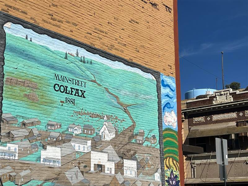 A mural titled “Main Street Colfax 1881” is painted on the side of a brick building. The top portion of a historic building and water tower are visible to the right of the mural, under a blue sky.