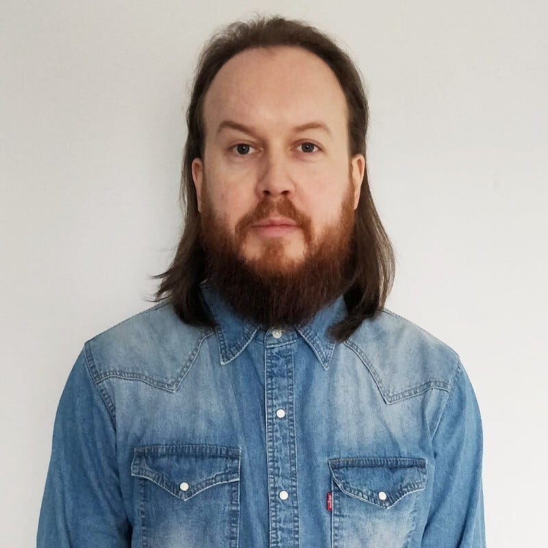 A picture of Mark, a white man with long brown hair and beard wearing a denim shirt.