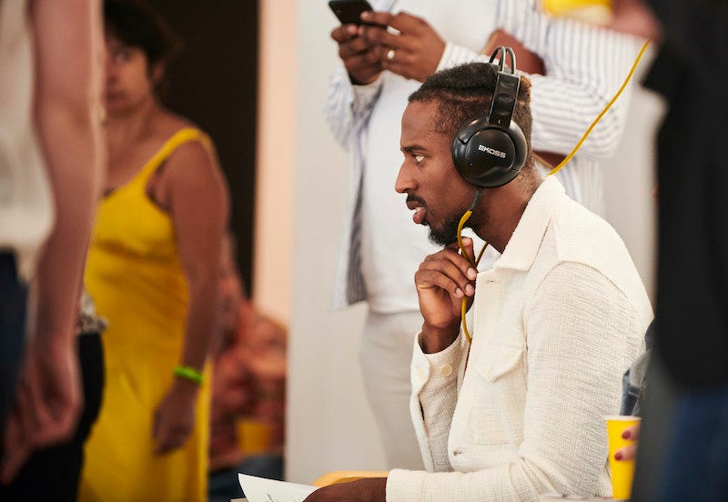 A black man sits with headphones on his head and his hand resting on his chin.