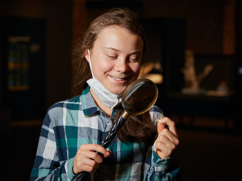 Climate activist Greta Thunberg stands smiling looking through a large magnifying glass at an unidentified small object pinched between her thumb and forefinger