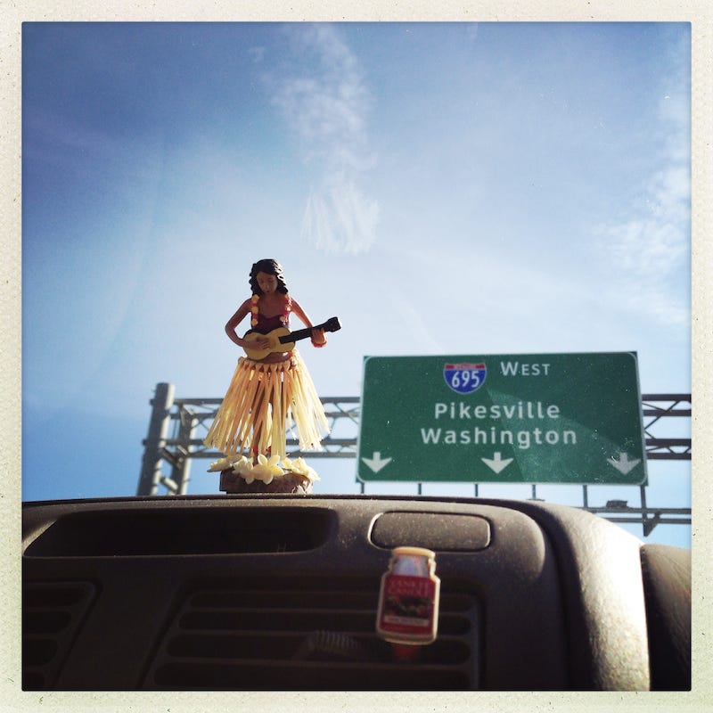 A hula girl figurine on a dusty dashboard with a highway sign in the background.