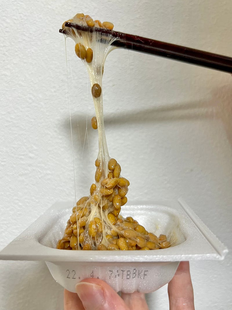 Natto fermented soybeans are held by chopstics above the pack, showing long strings of slime hanign down.