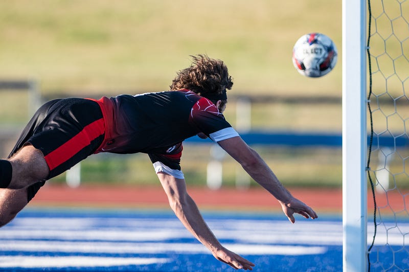 A man dives across screen with a football just to the right of his head, framed by a goal post