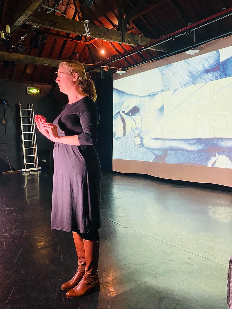 A BSL interpreter stands in front of a screen onto which there is a project black and white image of a blindfolded woman on her side.