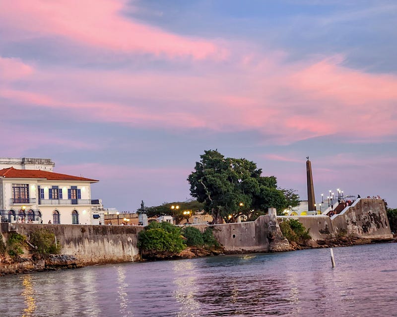 Pink and blue sunset sky over a peninsula with a pillar monument, trees, a lighted walkway, and antique-looking large white building.