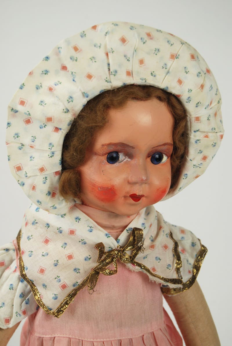 Textile doll filled with straw, head made of pressed cardboard. The doll is wearing a bonnet and a pink dress, has rosy chick and big eyes.