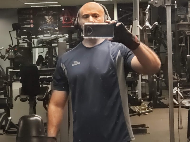 A bodybuilder takes a selfie at the gym, with weight-lifting equipment visible in the background