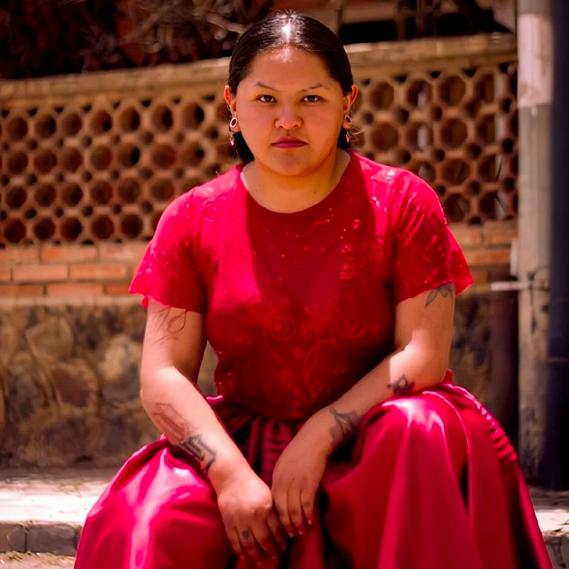 Alwa, a young brown skinned Bolivian woman with black hair pulled back neatly and parted in the middle. She is squatting in the street in an older neighborhood in a bright red silky thick dress making a stoic face. She has tattooed arms.