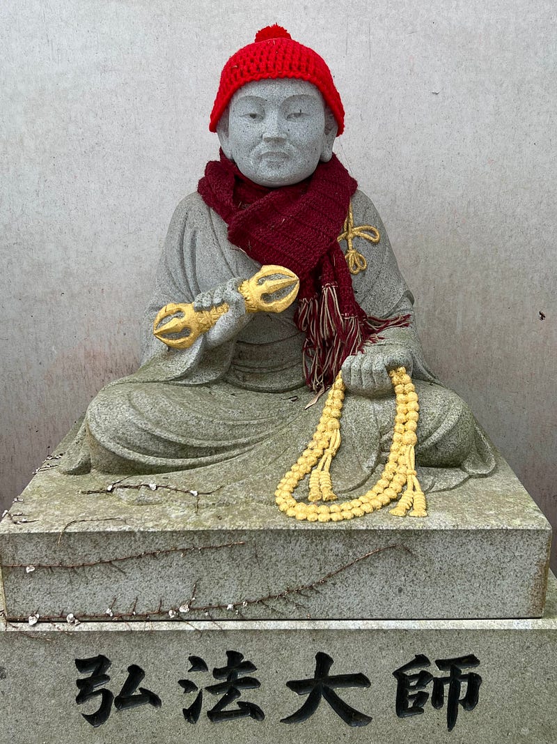 Stone statue with red cap, scarf, golden vajra, and prayer beads.