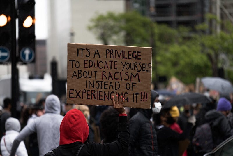 Picture of the backs of a crowd of people with someone holding up a piece of cardboard on which is written “Its a privilege to educate yourself about racism instead of experieicng it!!”