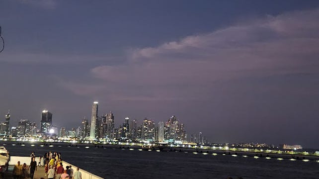 Lighted city skylines at desk across a dark ocean front and darky blue sky.