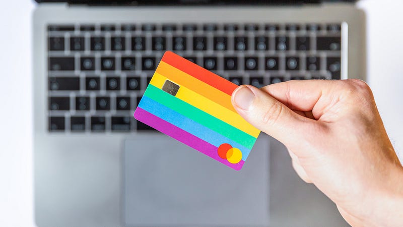 colourfully striped bank card with a mastercard-like logo held by a hand, above the keyboard of a silver apple-style laptop