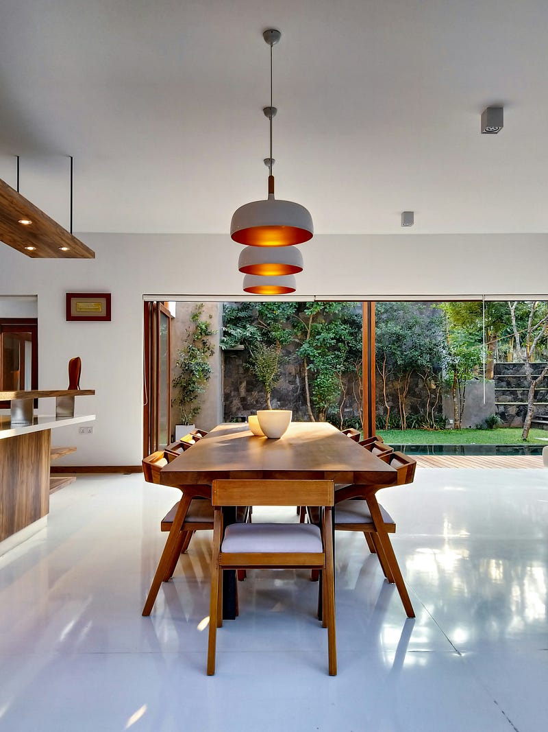 This modern dining area features a solid wooden dining table paired with sleek, minimalist chairs, creating a warm and inviting atmosphere. Stylish pendant lights with soft ambient glow enhance the dining experience, while floor-to-ceiling glass doors open to a lush green garden, bringing in natural light and freshness. Clean flooring, natural materials, and seamless indoor-outdoor connection make this dining space perfect for contemporary residential interiors.
