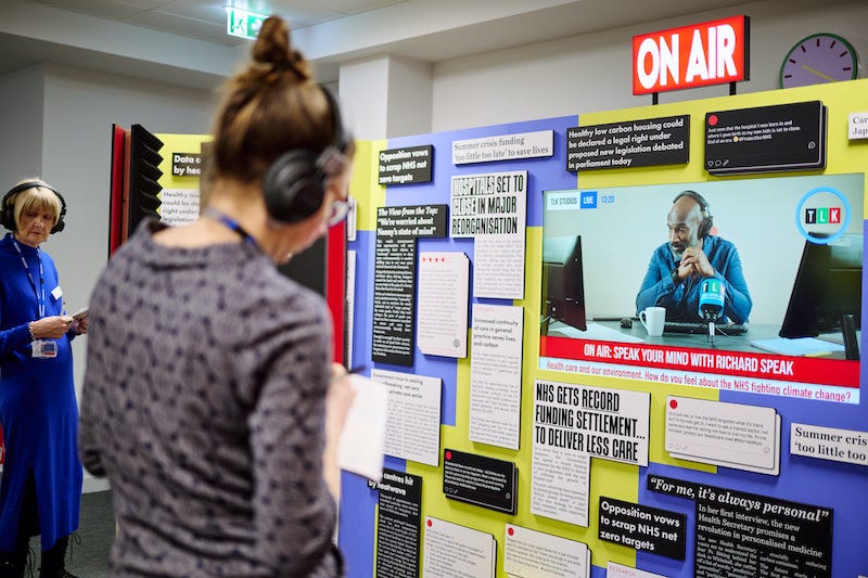 Two white women stand wearing headphones in front of a large board covered in news headlines and articles, and a screen displaying a news report.