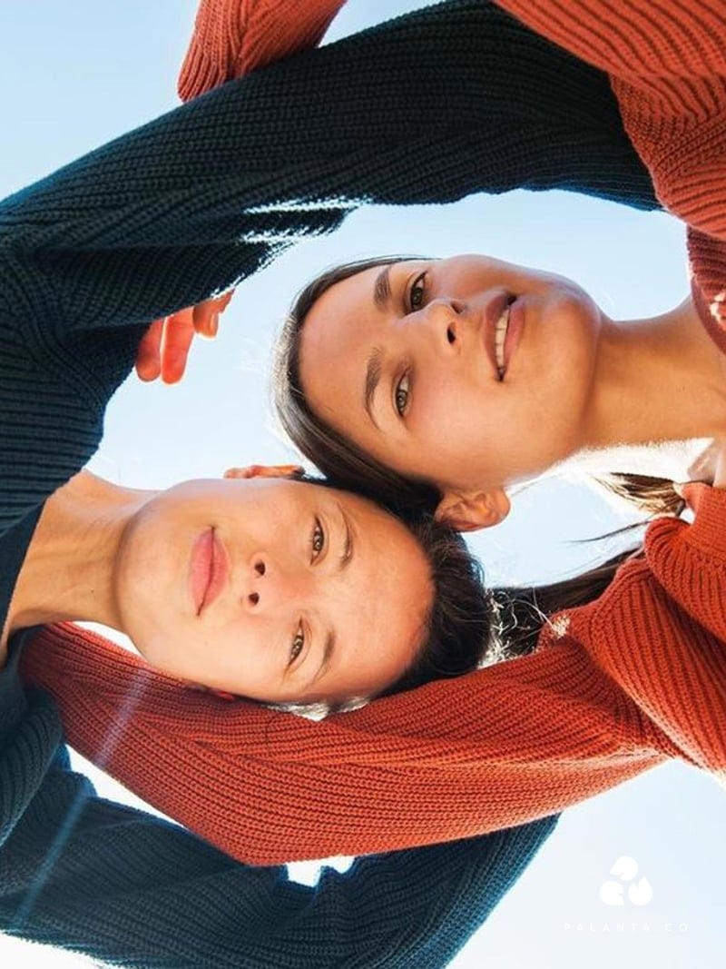 A photograph of two women. The camera is positioned on the ground as the two women lean over it from opposite sides, their arms reaching over each other’s shoulders. They wear warm jumpers, one black, one orange.