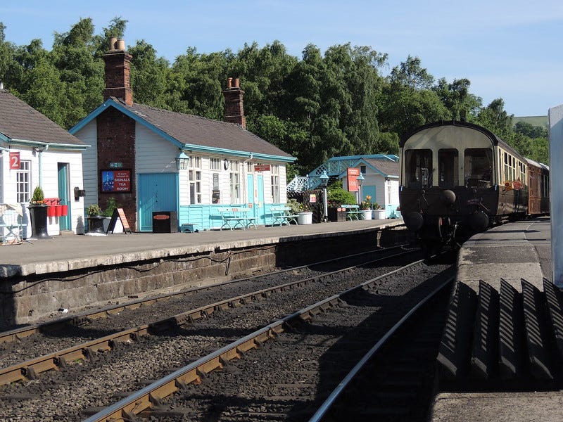 An old brown train pulling away from a small train station by a baby blue building