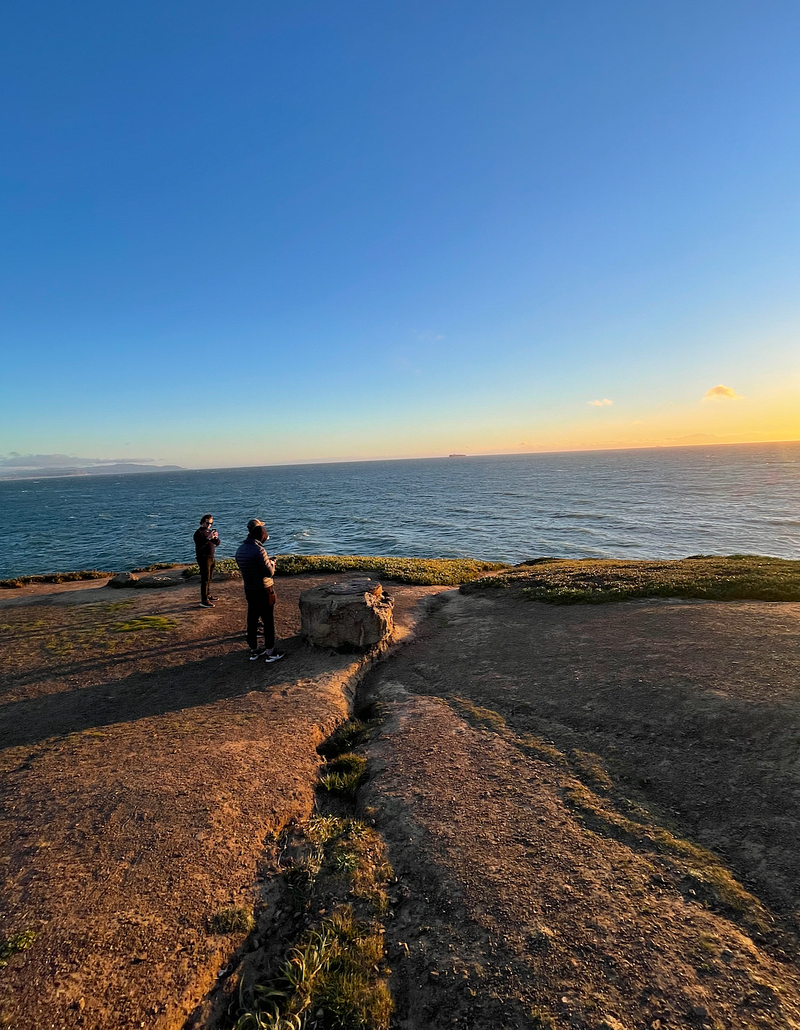 My friends overlooking the beautiful California coast. They’re on a rock cliff staring at the sunset starting to overtake the ocean.