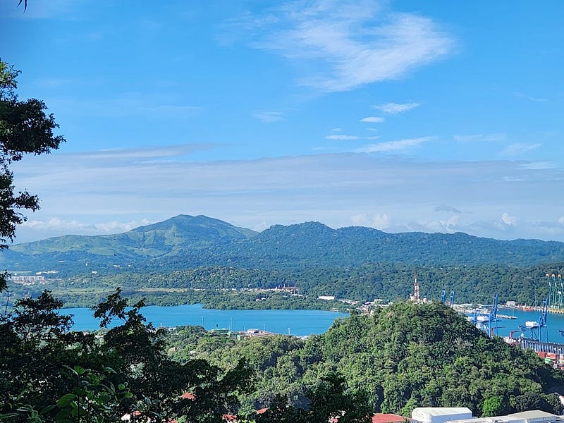 In the foreground of forested hilltops, in the midground, a bright blue canal, and in the background, green mountains line the horizon below a vivid, blue sky with wispy, white clouds.