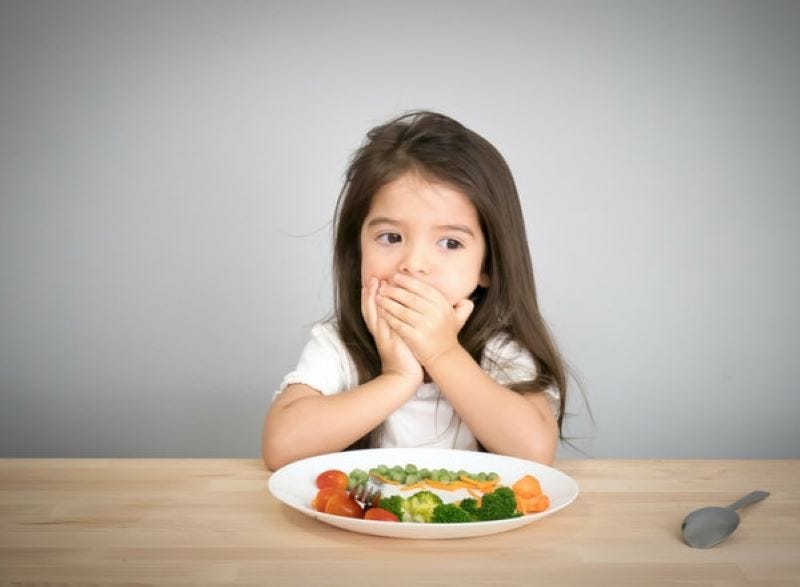A young girl sitting at a dining table with food in front of her, covering her mouth or we call it the Mouth Shut Movement (MSM), with her hands despite the food being served