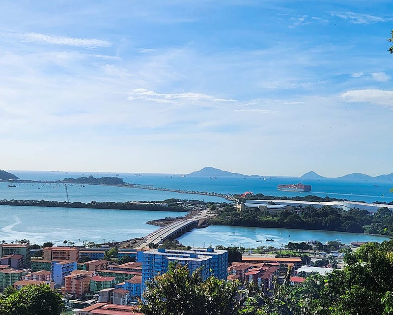 Landscape from above, including a blue sky with white wispy clouds, a bright blue ocean dotted with mountain islands, a bridge, and buildings.